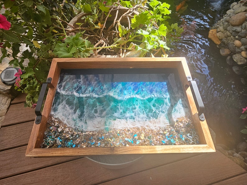 May include: A wooden serving tray with black metal handles features a resin ocean wave design with blue, green, and white colours. The tray is filled with small, iridescent blue and gold glitter.
