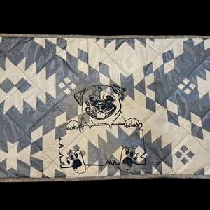 May include: A rectangular dog bed with a geometric pattern in white and blue. The center features a smiling pug illustration holding a sign with paw prints. The bed has a patterned border.