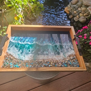 May include: A wooden serving tray with a resin inlay depicting a beach scene with blue and white waves crashing on a sandy shore. The resin is embedded with small blue and gold glitter.