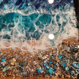 May include: A close-up of a resin art piece depicting a beach scene with turquoise blue water, white foam, and a sandy shore. The shore is decorated with small, iridescent glass pieces and a turquoise sea turtle figurine.