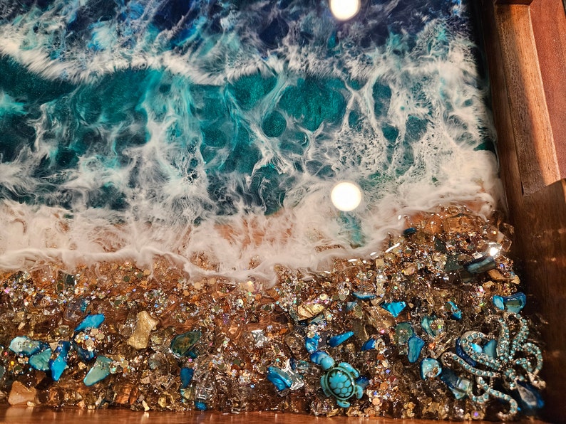 May include: A close-up of a resin art piece depicting a beach scene with turquoise blue water, white foam, and a sandy shore. The shore is decorated with small, iridescent glass pieces and a turquoise sea turtle figurine.
