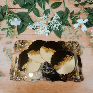 May include: A black and gold resin tray with four coasters. The tray has a unique, free-flowing design and a clear acrylic handle. The coasters are round and have a black and gold marbled design.