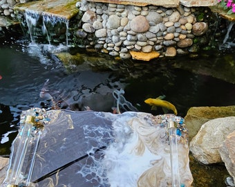 May include: A decorative resin tray with a unique design. The tray features a marbled pattern in shades of gray, white, and gold. It has clear acrylic handles with gold accents and is embellished with blue and silver crystals. The tray is ideal for serving or display.