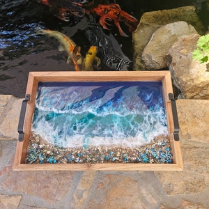 May include: A wooden tray with a resin ocean scene. The tray features a blue and green ocean with white foamy waves and a sandy beach with blue and green glitter. The tray has black metal handles.