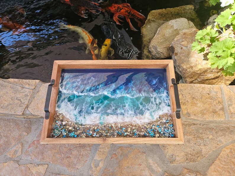 May include: A wooden tray with a resin ocean scene. The tray features a blue and green ocean with white foamy waves and a sandy beach with blue and green glitter. The tray has black metal handles.