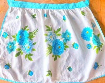 May include: A white waist apron featuring a bright turquoise blue floral pattern with green leaves. The apron has a turquoise blue waistband and matching trim along the edges. The fabric is gathered at the waist, creating a pleated effect.