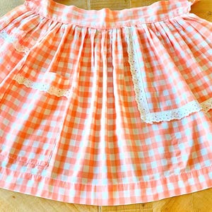 May include: A waist apron featuring a peach and white gingham check pattern. The apron has a gathered skirt, a wide waistband, and two front pockets. Each pocket is accented with a white eyelet lace trim along the top and side edges.