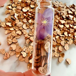 May include: A clear glass vial with a cork stopper containing a single purple flower and numerous small wooden heart-shaped pieces, each engraved with the word love, surrounded by a pile of identical wooden hearts.