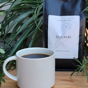 May include: A white ceramic mug filled with dark coffee sits on a wooden table next to a black bag of coffee beans. The bag has a white label with the text "Spice Theory Coffee Company" and an illustration of an owl with the text "Dark Specialty Blend".