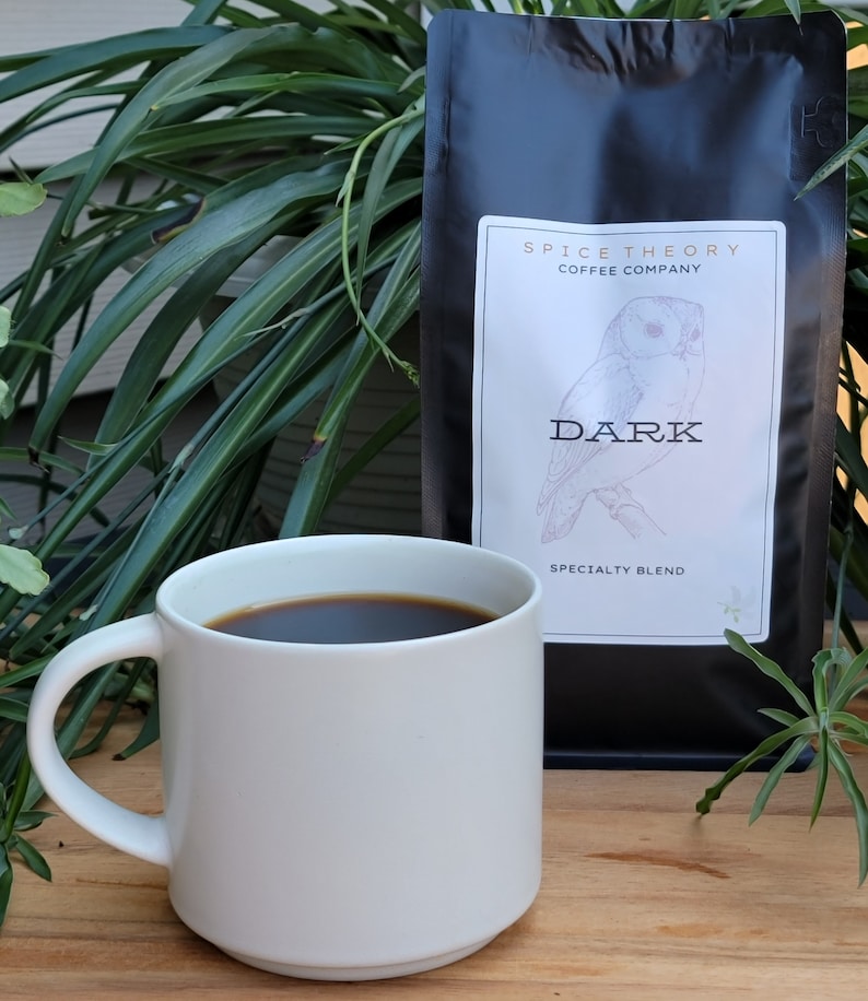May include: A white ceramic mug filled with dark coffee sits on a wooden table next to a black bag of coffee beans. The bag has a white label with the text "Spice Theory Coffee Company" and an illustration of an owl with the text "Dark Specialty Blend".