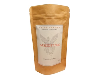 Puede incluir: Una bolsa de papel marrón con café y una etiqueta blanca. La etiqueta dice "SPICE THEORY COFFEE COMPANY" sobre una ilustración detallada de un pájaro. Debajo de la ilustración, la palabra "MEDIUM" está en rojo. "SPECIALTY BLEND" está impreso en la parte inferior.