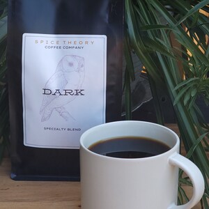 May include: A black bag of coffee beans with a white label that says "Spice Theory Coffee Company" and "Dark Specialty Blend". The label also features a drawing of an owl. A white mug with a dark brown coffee inside is sitting on a wooden table.