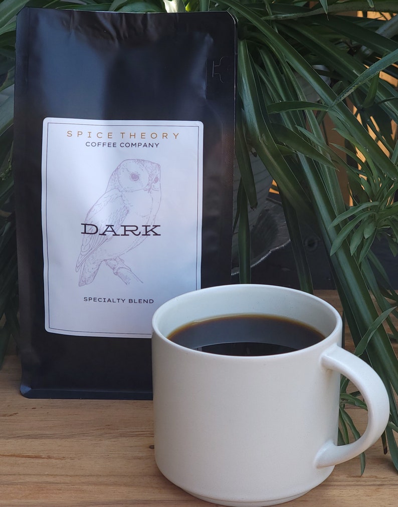 May include: A black bag of coffee beans with a white label that says "Spice Theory Coffee Company" and "Dark Specialty Blend". The label also features a drawing of an owl. A white mug with a dark brown coffee inside is sitting on a wooden table.