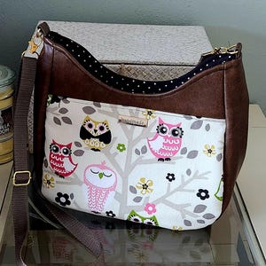 May include: A brown leather and fabric shoulder bag with a white owl print. The bag has a long brown strap and a flap closure. The owl print features pink, white, and gray owls on a white background with brown branches.