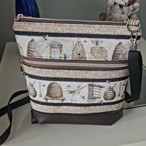 May include: A brown and black fabric crossbody bag with a zipper closure and a strap. The bag has a bee and honeycomb theme with a brown, black, and white pattern. The bag has two compartments.