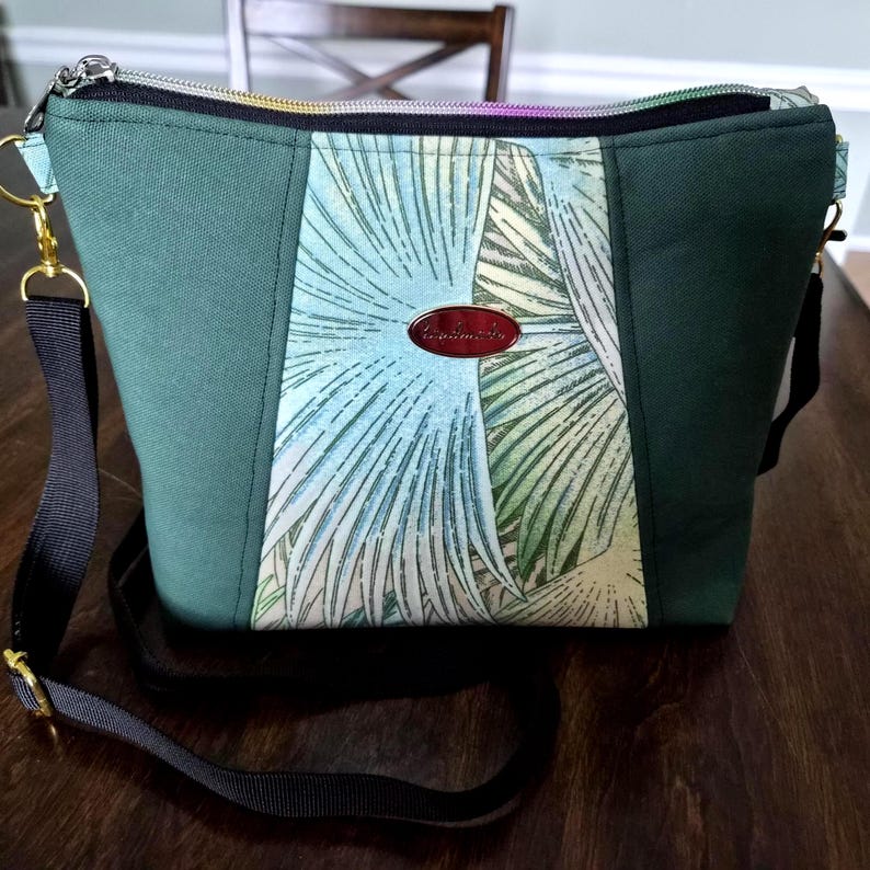May include: A green crossbody bag with a central panel featuring a tropical leaf pattern in shades of blue, green, and cream. The bag has a black strap, gold-colored hardware, and a zipper closure. A small, oval, red tag is attached to the front.