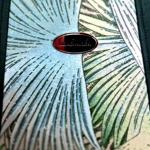 May include: Close-up of a fabric with a tropical leaf pattern in shades of blue, green, and cream. A small, oval, copper-colored tag with the word "handmade" is attached to the fabric.