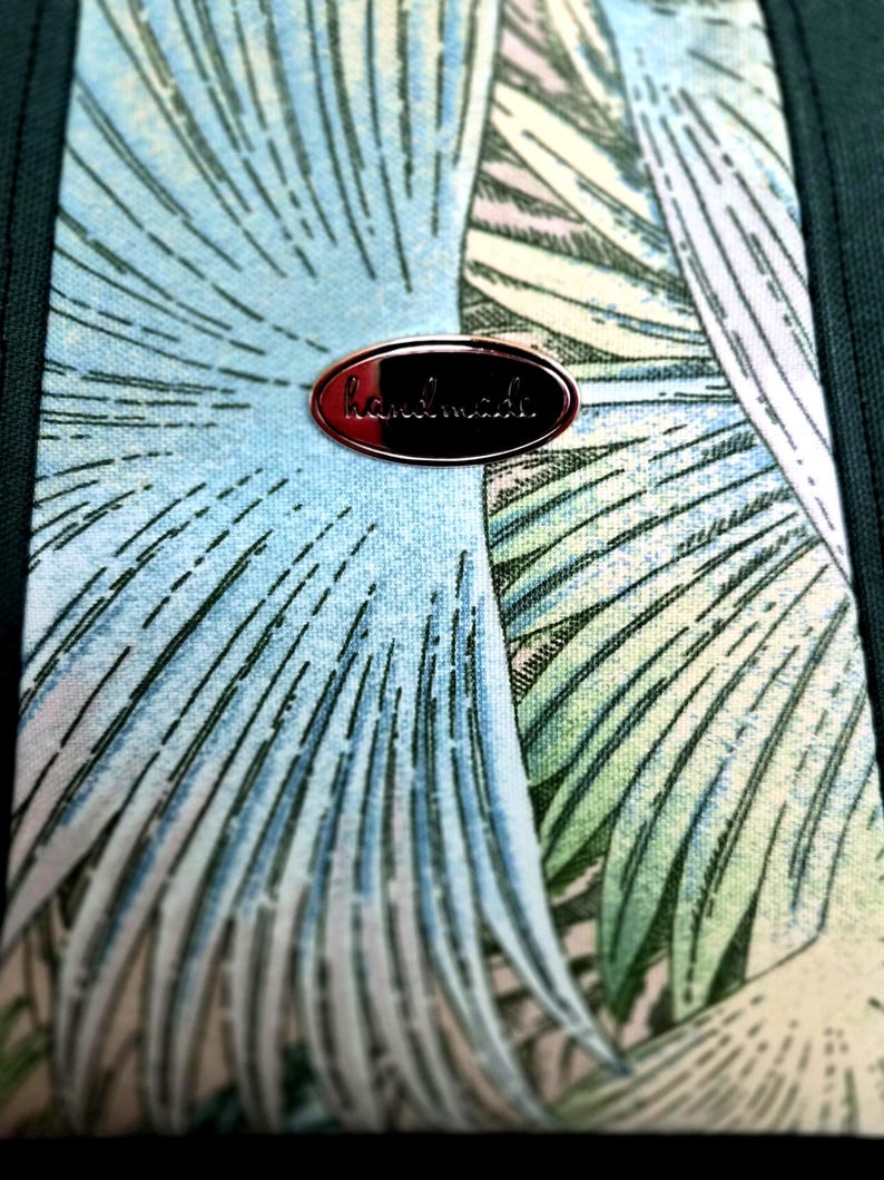 May include: Close-up of a fabric with a tropical leaf pattern in shades of blue, green, and cream. A small, oval, copper-colored tag with the word "handmade" is attached to the fabric.
