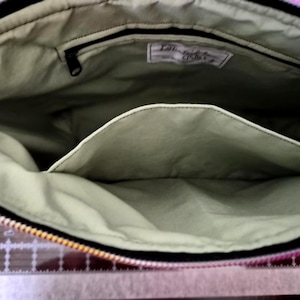May include: Open view of a fabric bag interior. The bag has a light green lining with a black zipper and a pocket. A small tag is visible inside the bag. The bag's exterior is black with a colorful pattern.
