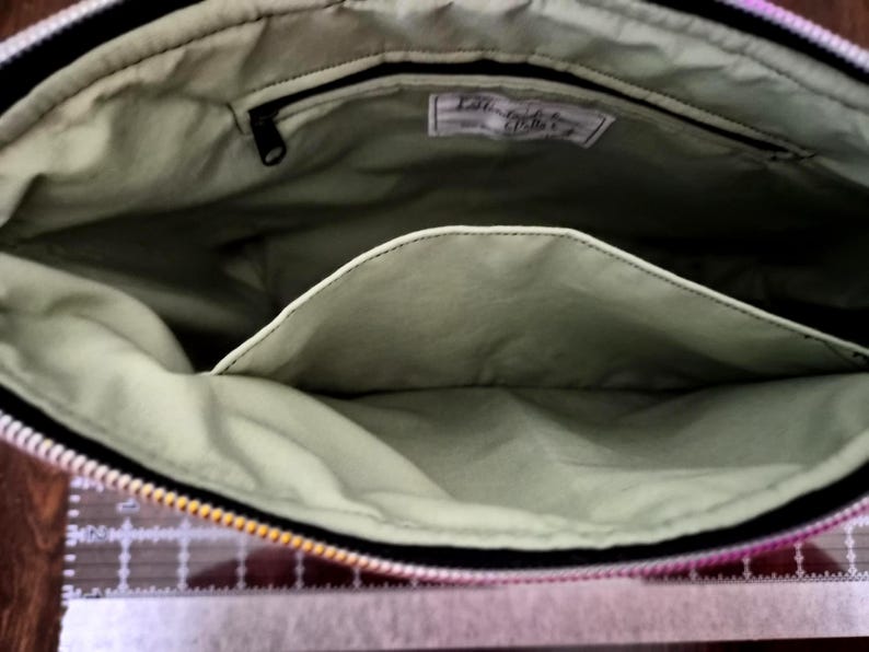 May include: Open view of a fabric bag interior. The bag has a light green lining with a black zipper and a pocket. A small tag is visible inside the bag. The bag's exterior is black with a colorful pattern.