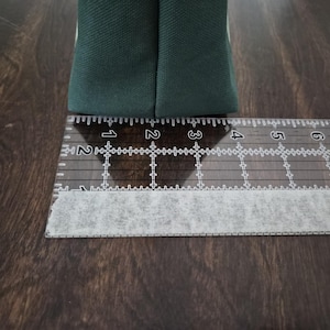 May include: A close-up of a dark green fabric item, possibly a bag or pouch, next to a clear ruler. The ruler shows measurements in inches, with the fabric item spanning approximately 2 inches.