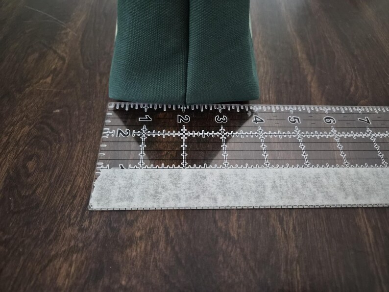 May include: A close-up of a dark green fabric item, possibly a bag or pouch, next to a clear ruler. The ruler shows measurements in inches, with the fabric item spanning approximately 2 inches.