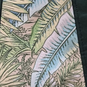 May include: Close-up of a fabric with a tropical leaf pattern. The design features large, light green, and blue leaves against a beige and pink background. Dark green fabric borders the patterned section, creating a frame.