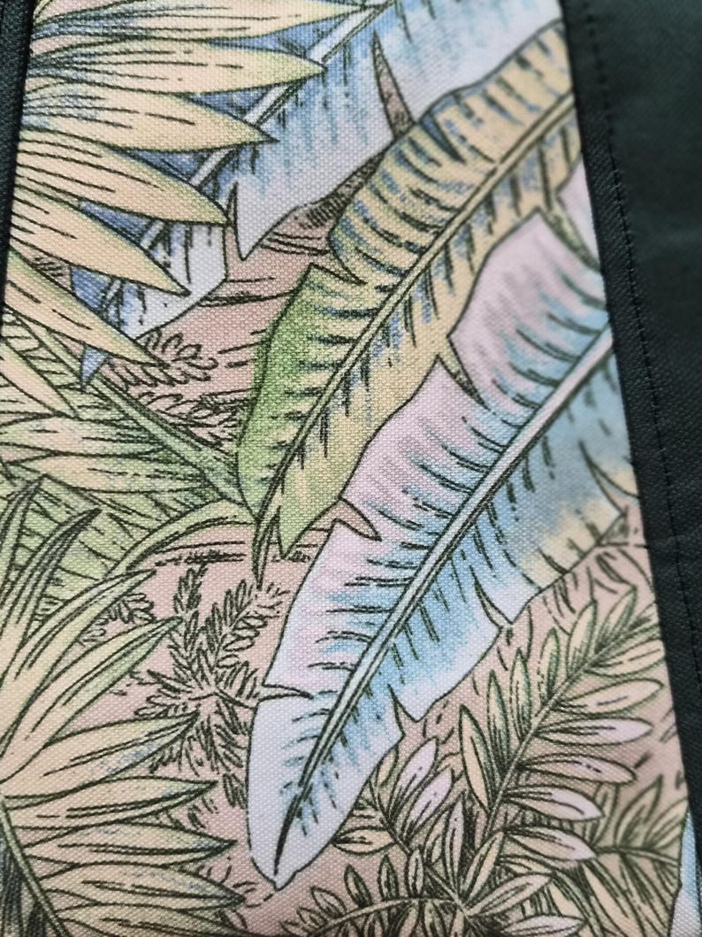 May include: Close-up of a fabric with a tropical leaf pattern. The design features large, light green, and blue leaves against a beige and pink background. Dark green fabric borders the patterned section, creating a frame.