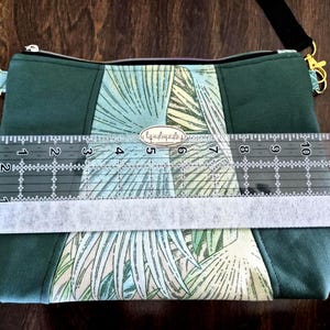 May include: Green crossbody bag with a palm leaf pattern and a black adjustable strap. The bag is approximately 12 inches wide. The bag has a zipper closure and gold-colored hardware. The bag has a small tag that says "handmade".