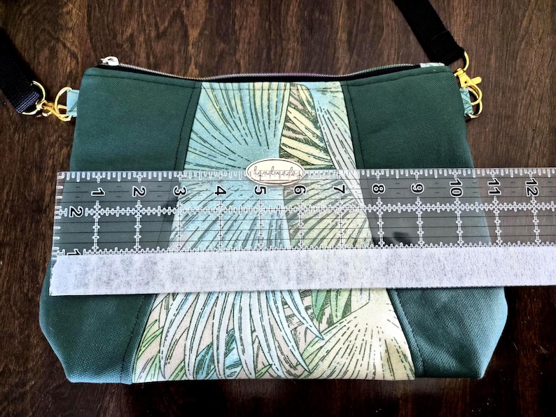 May include: Green crossbody bag with a palm leaf pattern and a black adjustable strap. The bag is approximately 12 inches wide. The bag has a zipper closure and gold-colored hardware. The bag has a small tag that says "handmade".