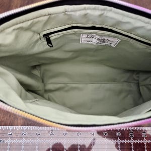 May include: Open view of a handmade zippered bag with a light green interior. The bag's interior features a small zippered pocket and a label with text. The exterior has a rainbow gradient design. A ruler is at the bottom.