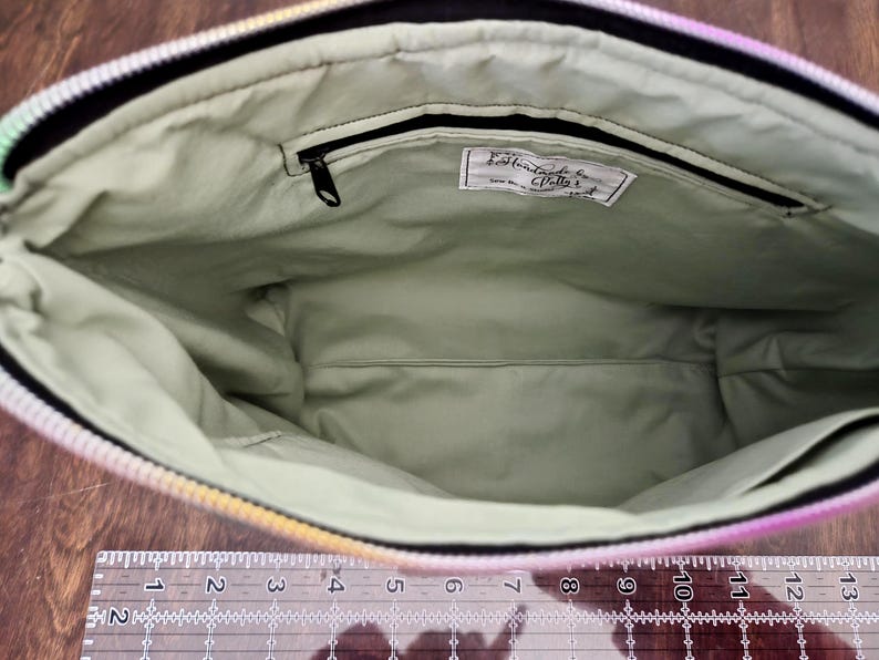 May include: Open view of a handmade zippered bag with a light green interior. The bag's interior features a small zippered pocket and a label with text. The exterior has a rainbow gradient design. A ruler is at the bottom.