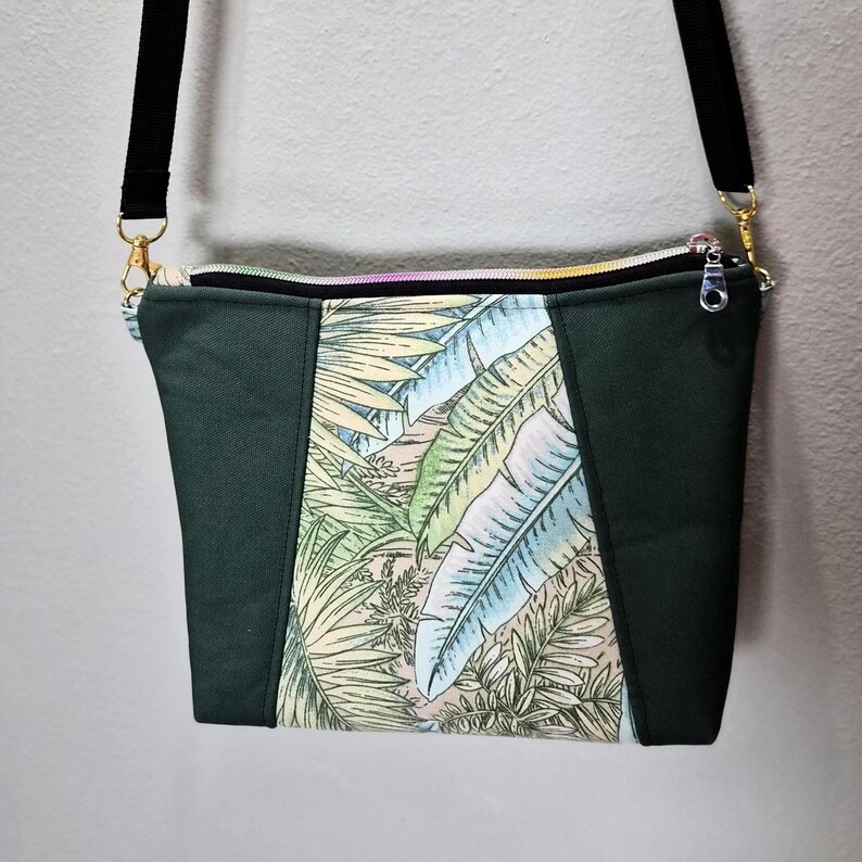 May include: A dark green crossbody bag with a black strap and gold-colored hardware. The center panel features a tropical leaf print in shades of green, blue, and beige. The bag has a zippered closure.