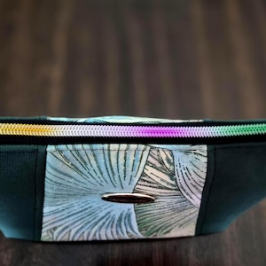 May include: A dark green cosmetic bag with a colorful zipper and a floral pattern. The bag has a silver-colored oval emblem on the front. The zipper has sections of gold, purple, and green.