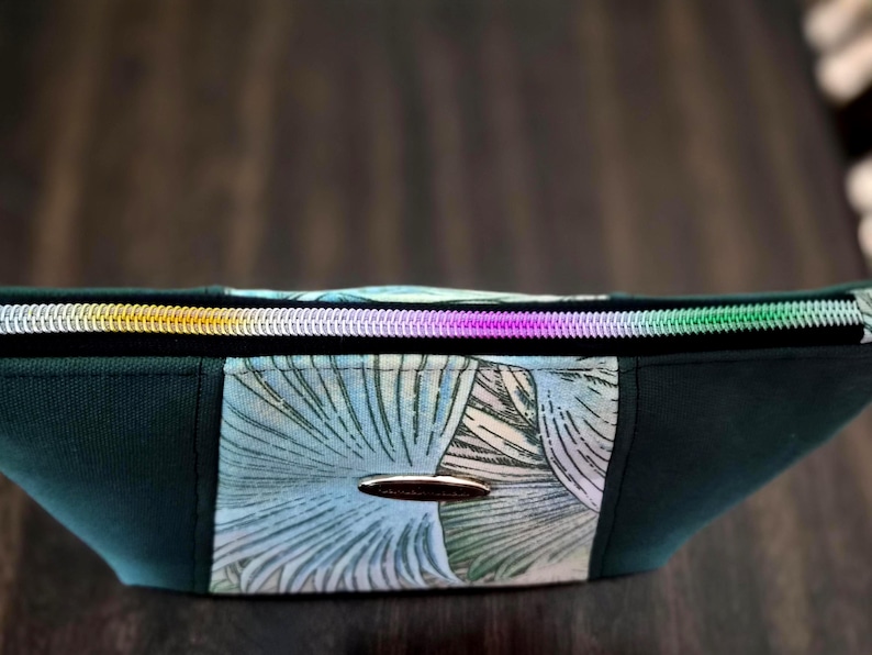 May include: A dark green cosmetic bag with a colorful zipper and a floral pattern. The bag has a silver-colored oval emblem on the front. The zipper has sections of gold, purple, and green.