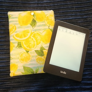 May include: A black Kindle e-reader next to a fabric case. The case features a lemon pattern with yellow lemons, green leaves, and white and light blue stripes. The Kindle screen is white.