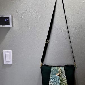 May include: A dark green crossbody bag with a black adjustable strap. The bag features a central panel with a palm leaf design in shades of green and blue. Gold-colored hardware accents the bag.