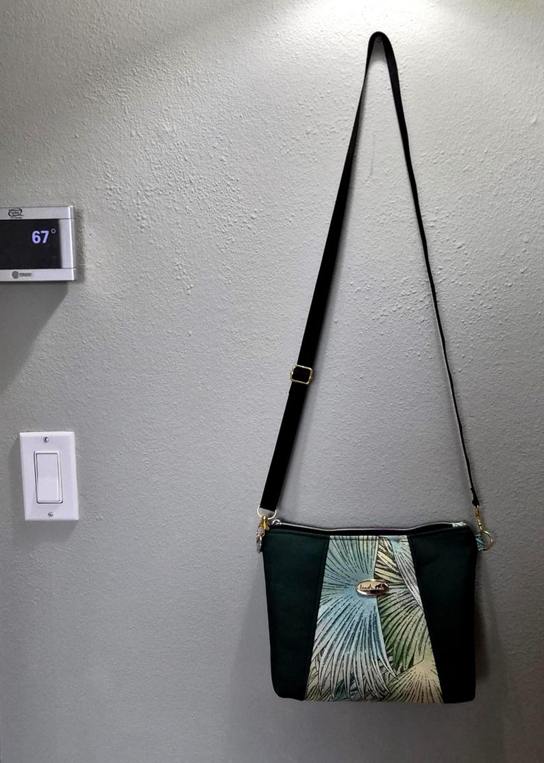 May include: A dark green crossbody bag with a black adjustable strap. The bag features a central panel with a palm leaf design in shades of green and blue. Gold-colored hardware accents the bag.