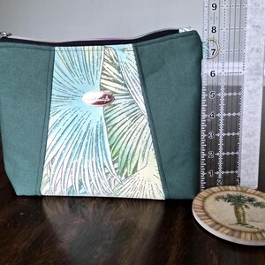 May include: A rectangular green and patterned fabric pouch with a black zipper and a small metal ring. The center panel features a tropical leaf design in shades of blue, green, and white. A ruler is next to the pouch. A coaster with a palm tree design is also present.