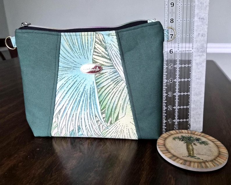 May include: A rectangular green and patterned fabric pouch with a black zipper and a small metal ring. The center panel features a tropical leaf design in shades of blue, green, and white. A ruler is next to the pouch. A coaster with a palm tree design is also present.