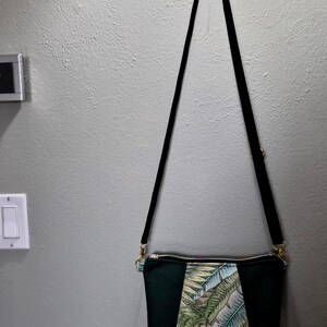 May include: A black crossbody bag with a long, black strap. The bag features a central panel with a tropical leaf pattern in shades of green, blue, and beige, framed by solid black panels. Gold-toned hardware accents the zipper and strap attachments.