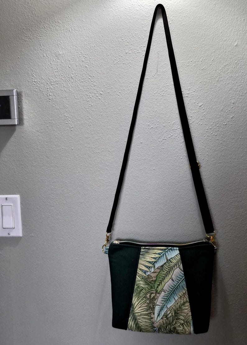 May include: A black crossbody bag with a long, black strap. The bag features a central panel with a tropical leaf pattern in shades of green, blue, and beige, framed by solid black panels. Gold-toned hardware accents the zipper and strap attachments.