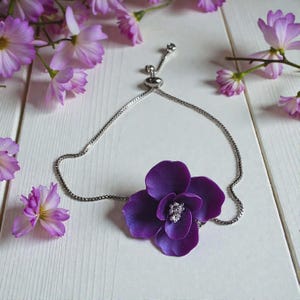 May include: A silver chain bracelet featuring a large, deep purple flower with a sparkling center. The flower is the focal point, set against a white wooden surface, with delicate purple flowers scattered around.
