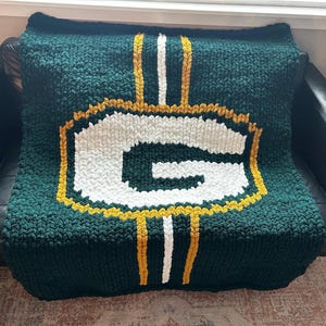 May include: A handmade, chunky knit blanket in dark green, featuring a large white letter "G" outlined in gold and a green border. Vertical stripes in white and gold accent the design. The blanket is folded over a black surface.