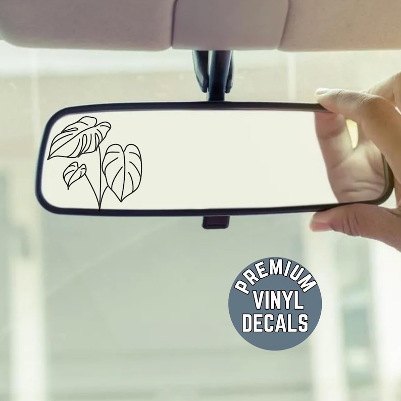 May include: A car's rearview mirror with a black outline of a plant design on the glass. The mirror is being held by a hand. A blue circle with the text "PREMIUM VINYL DECALS" is in the lower right corner.