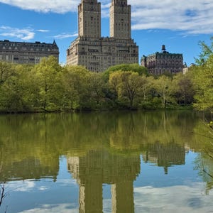 May include: A scenic view of a lake reflecting the image of a tall, ornate building with two towers. The building is surrounded by lush green trees and a bright blue sky with scattered clouds. The water's surface mirrors the architecture.
