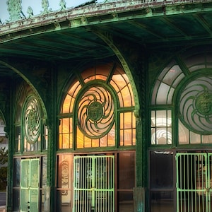 May include: An exterior view of a building with elaborate green metalwork. The structure has arched windows with decorative spiral designs and green metal gates. The roof has detailed embellishments. The windows have an orange glow.