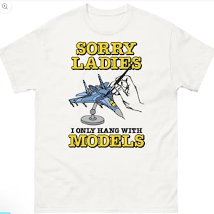 May include: White t-shirt with the text "SORRY LADIES I ONLY HANG WITH MODELS" in yellow and black. The graphic features a blue model airplane being painted by a hand. A humorous design for model enthusiasts.