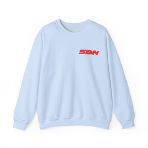 May include: A pale blue sweatshirt featuring the red letters "SDN" on the chest. This long-sleeved crew neck top is made of a soft material and is suitable for casual wear. The sweatshirt is a solid pale blue colour.