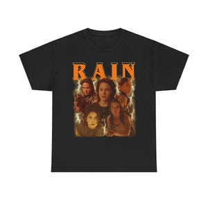 May include: Black t-shirt featuring a graphic print of a group of people from the film "Rain". The text "RAIN" is printed in orange above the image.